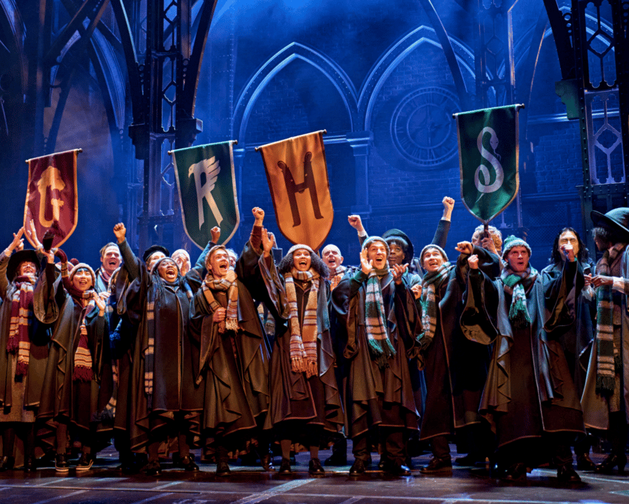 Cast on stage. Hogwarts students cheer in house hat & scarves with House banners behind.