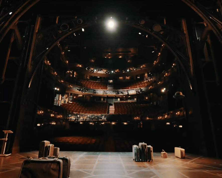 Cambridge Theatre seating seen from the stage. Suitcases are dotted around the stage as the camera looks to the tiered seats.